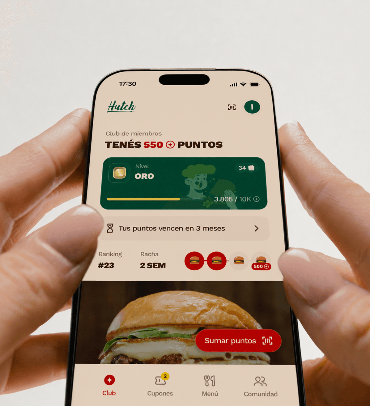 Dos manos sosteniendo un teléfono con la app del club de Hutch: 550 puntos, nivel Oro, la racha de dos semanas y el botón para sumar puntos