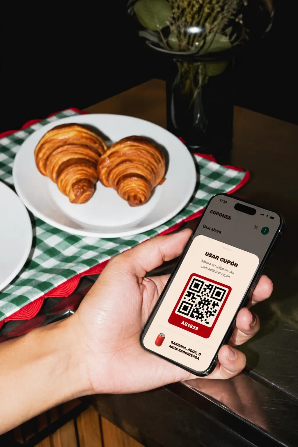 Una mano con el teléfono mostrando el cupón y su código QR para canjear, sobre una mesa de café con dos medialunas