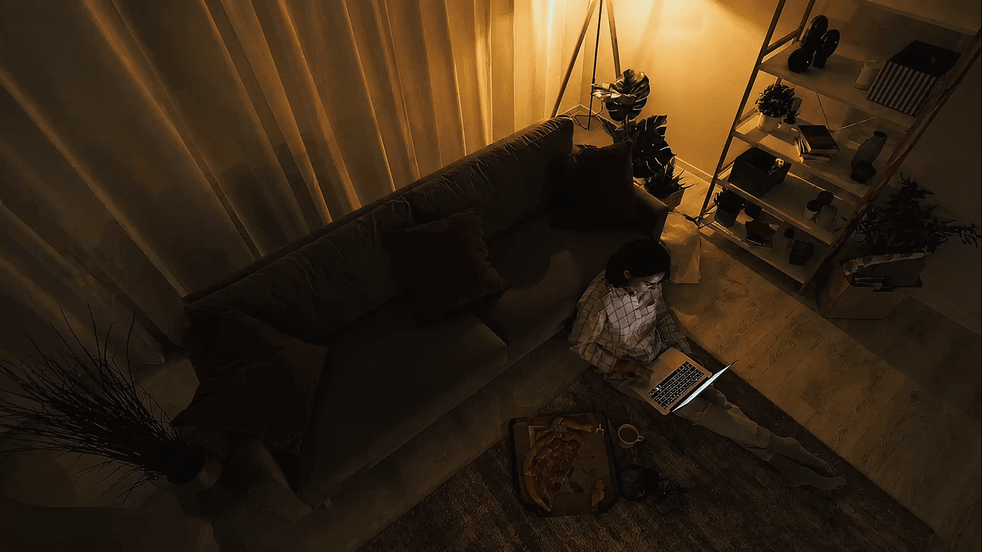Un living a oscuras visto desde arriba, con una persona sentada en el piso contra el sillón trabajando en su notebook, una pizza a medio comer al lado y una lámpara encendida al fondo