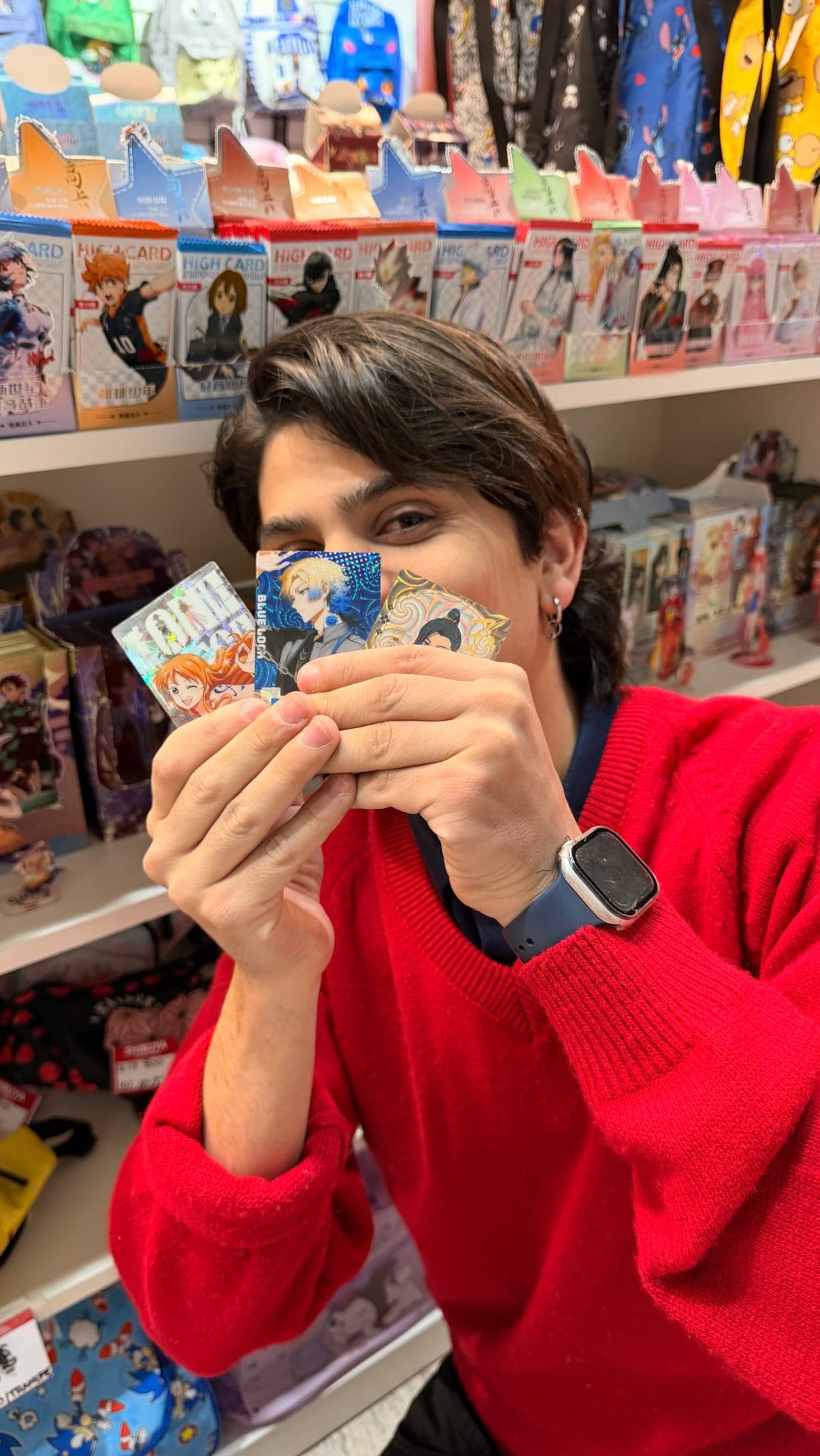 Shibuya photo: a person in a red hoodie holds a fan of cards in front of their face, with the store's shelves of figures and boxes behind them.