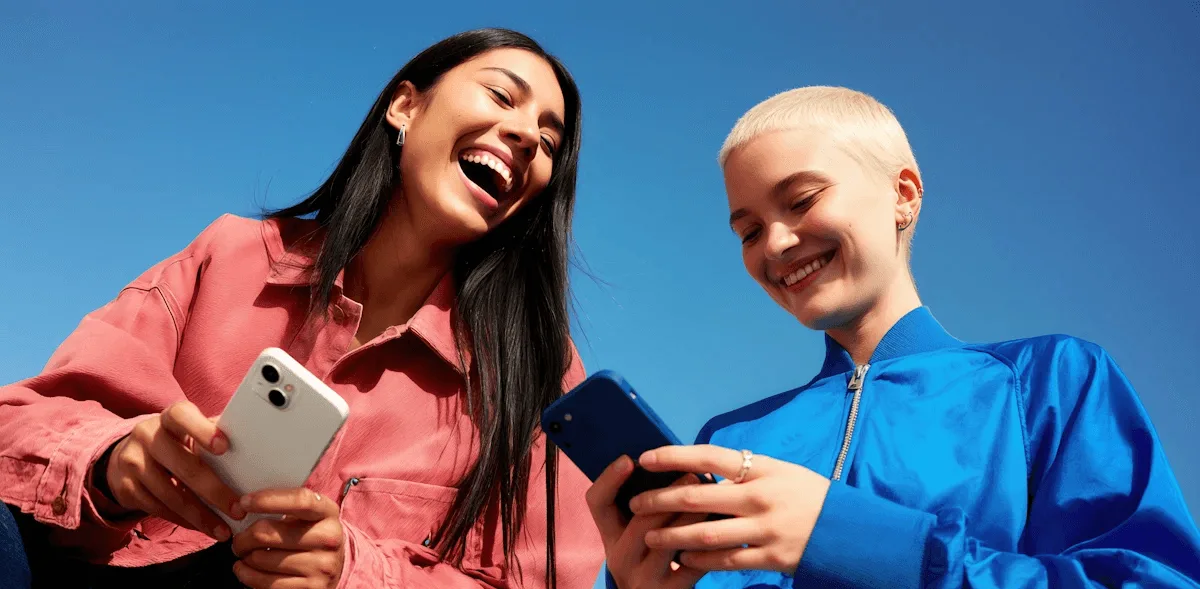 Dos amigas se ríen mirando un teléfono sobre un fondo azul