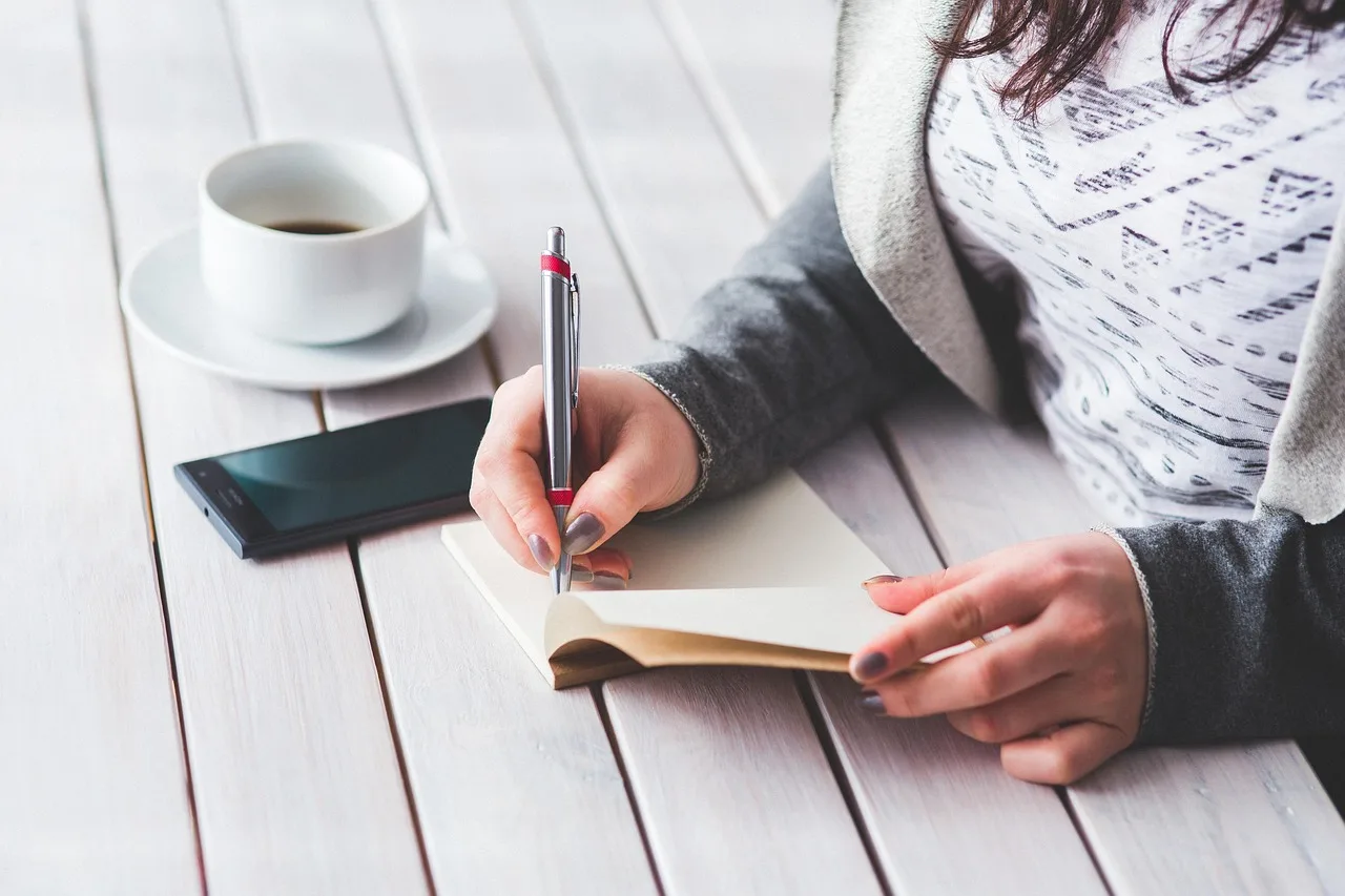 Una persona escribe en una libreta sobre una mesa de madera blanca, con un teléfono y una taza de café al lado