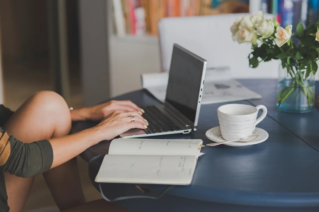 Una persona escribe en una computadora portátil sobre una mesa redonda con una taza de café y flores