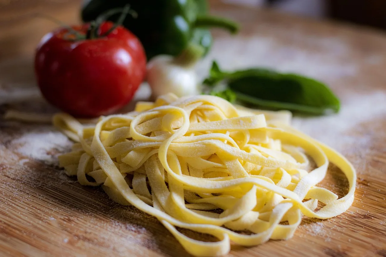 Un nido de pasta fresca sobre una tabla enharinada con un tomate y hojas verdes detrás