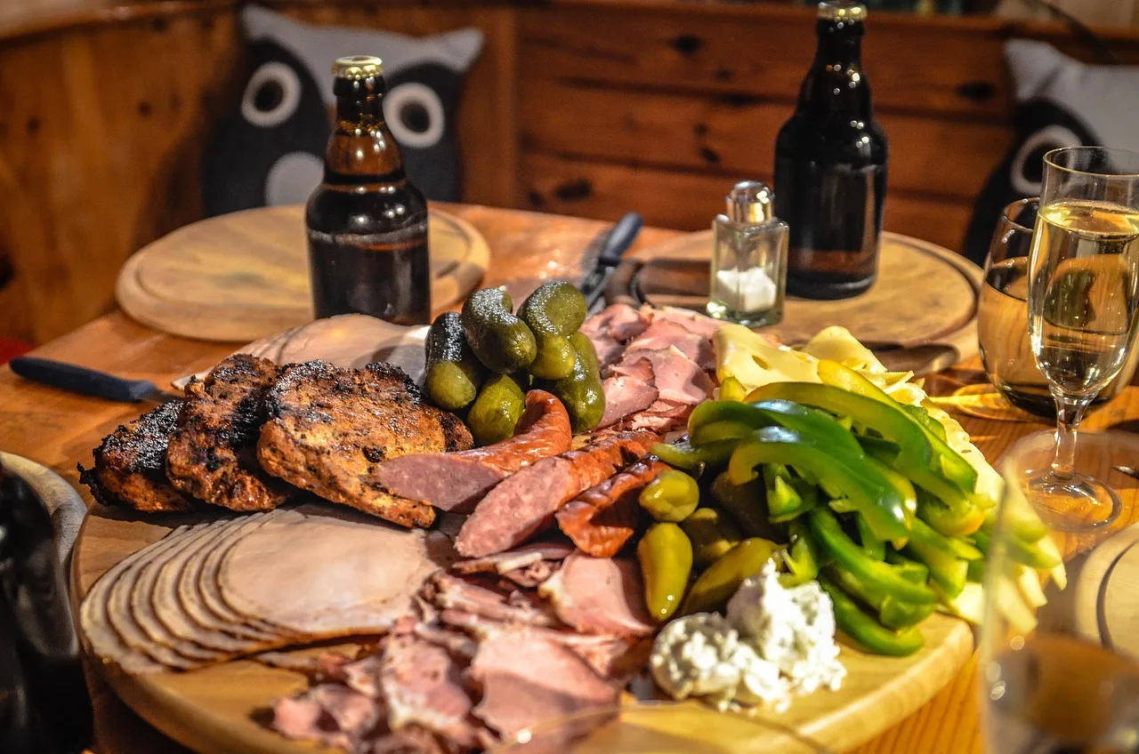Una tabla de madera con fiambres feteados, pepinillos y queso sobre una mesa con dos botellas oscuras