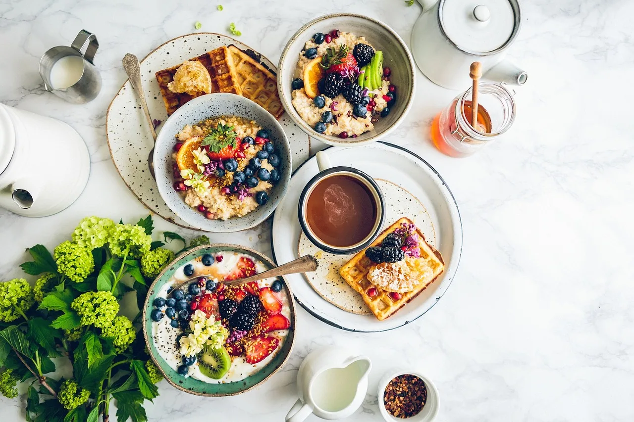 Una mesa de mármol vista desde arriba con waffles, bowls de avena con frutas, una taza de chocolate y miel