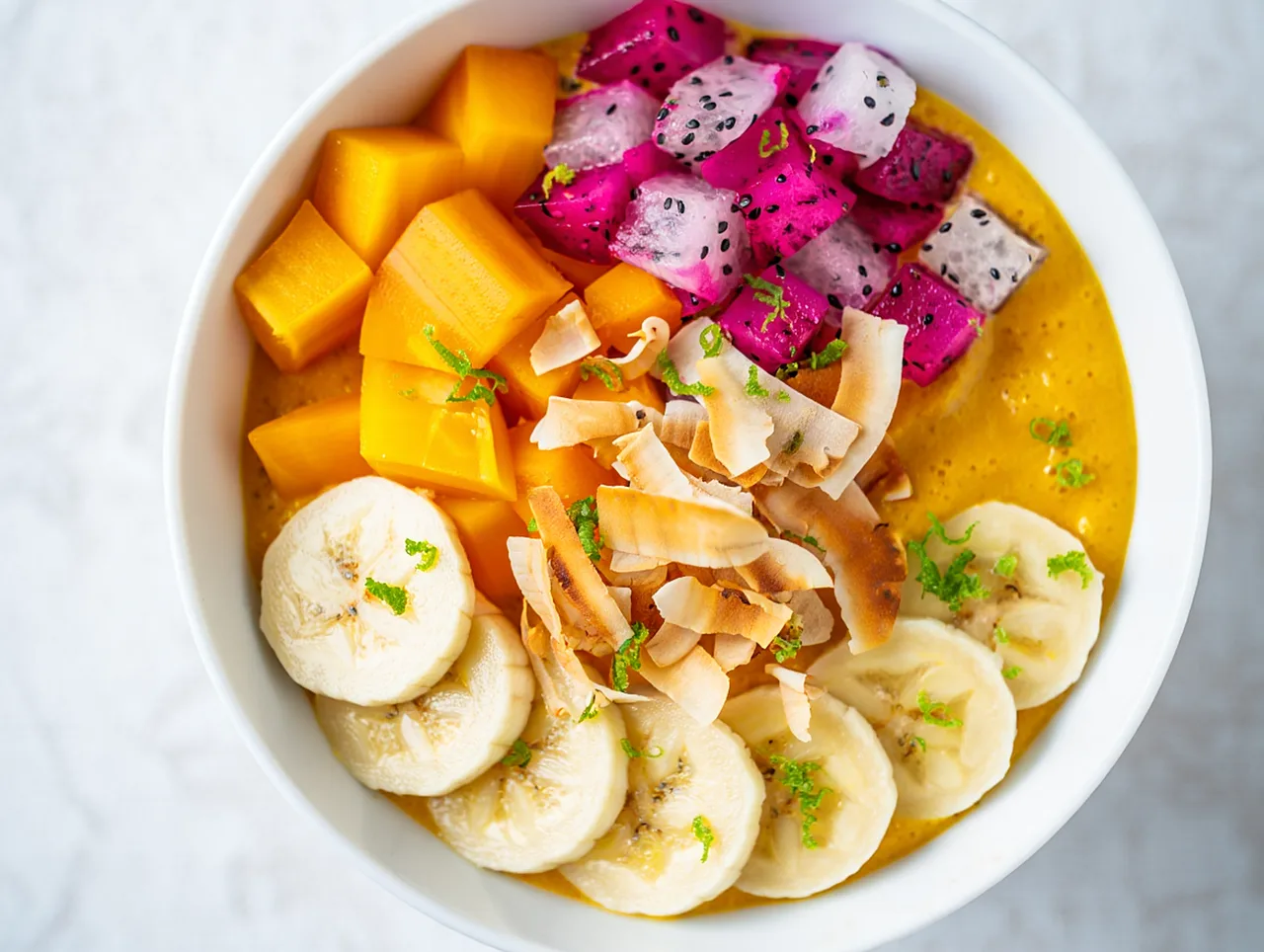 Un bowl de batido de mango con fruta picada, fruta del dragón y coco en láminas visto desde arriba sobre una mesa clara