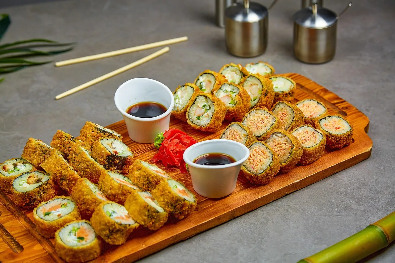 Una tabla de madera con rolls de sushi rebozados, dos cuencos de salsa de soja y palitos al costado
