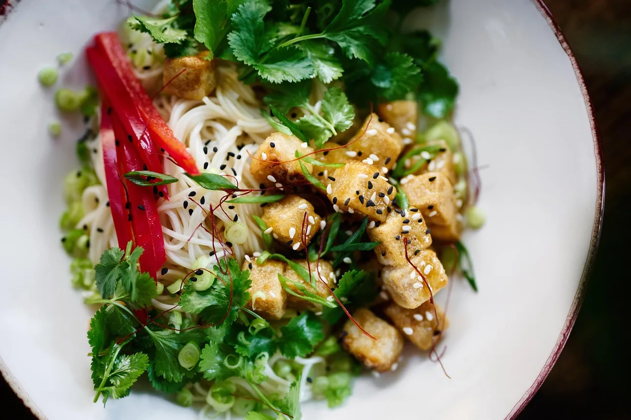 Un plato hondo con fideos, cubos de tofu dorado, hojas de cilantro y tiras de pimiento rojo