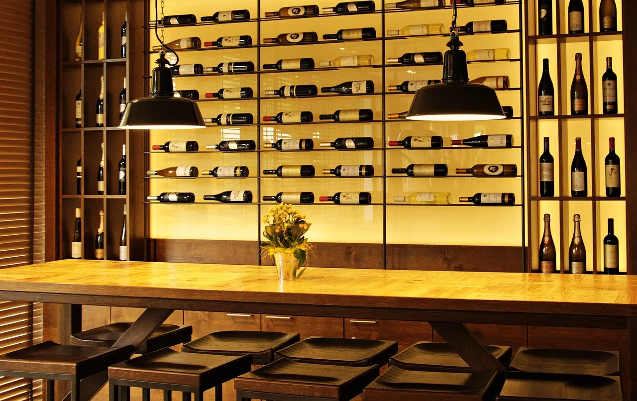 Una mesa larga de madera con bancos altos frente a una pared iluminada llena de botellas de vino acostadas