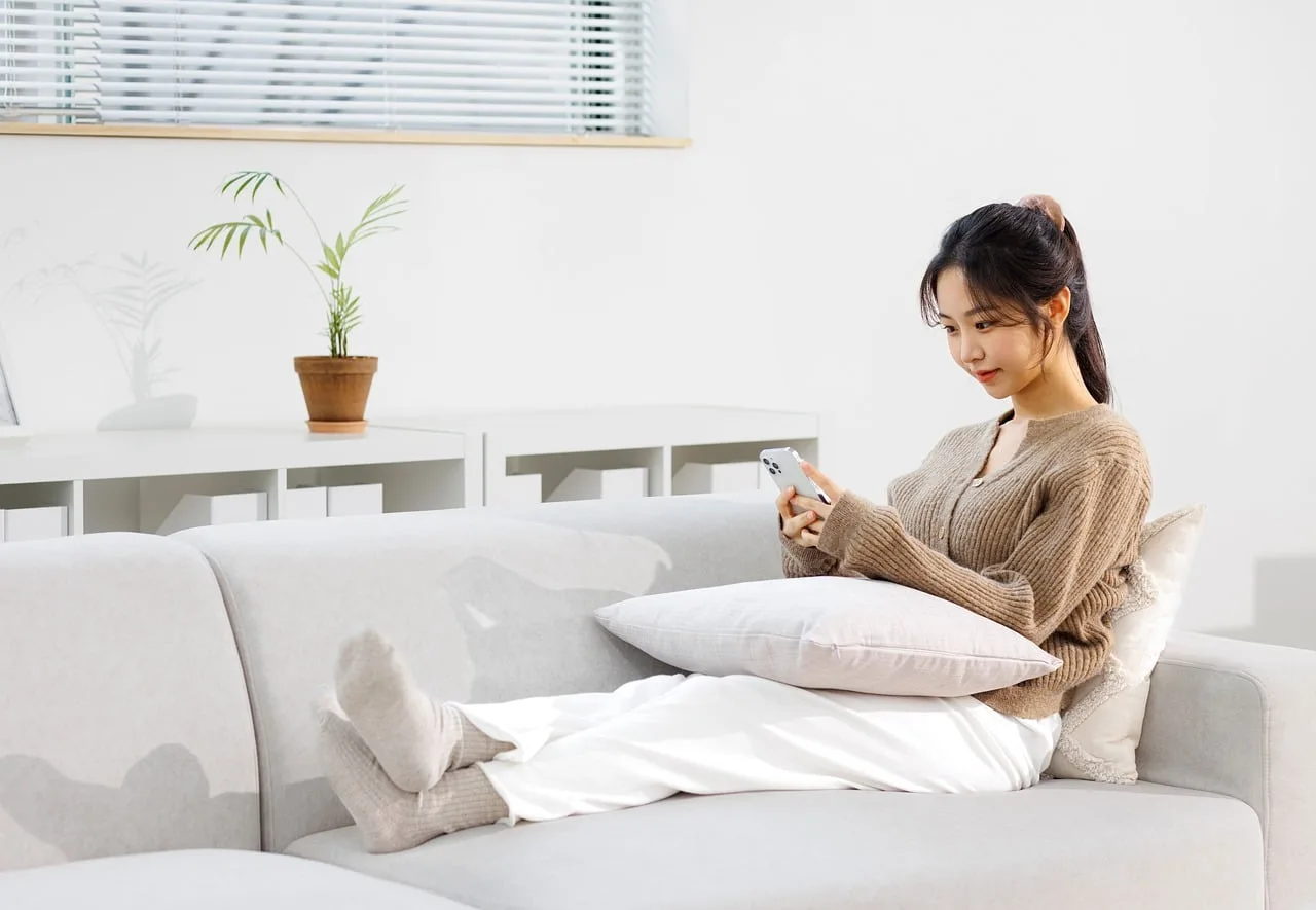 Una mujer sentada en un sillón blanco mira su teléfono en un living luminoso con una planta