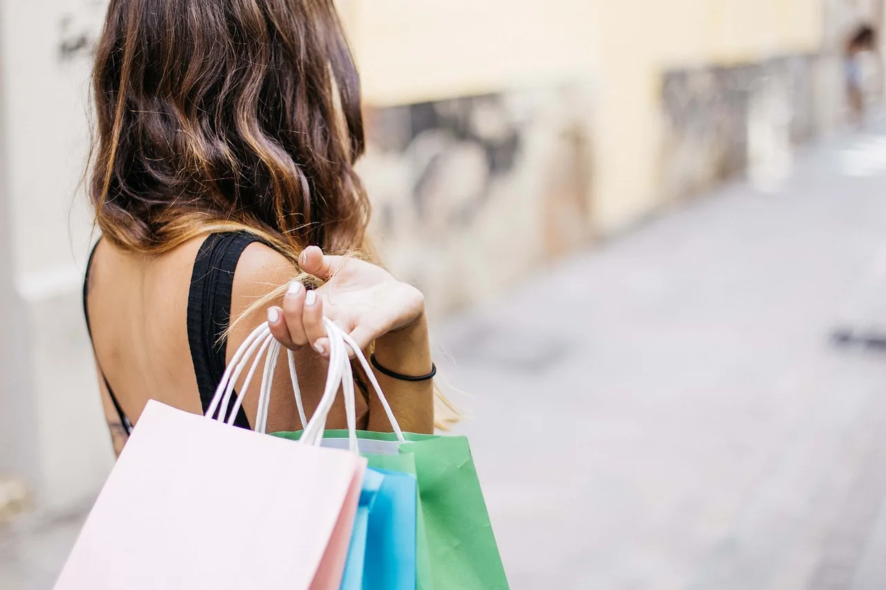 Una mujer de espaldas camina por una calle con varias bolsas de compras de colores pastel en la mano