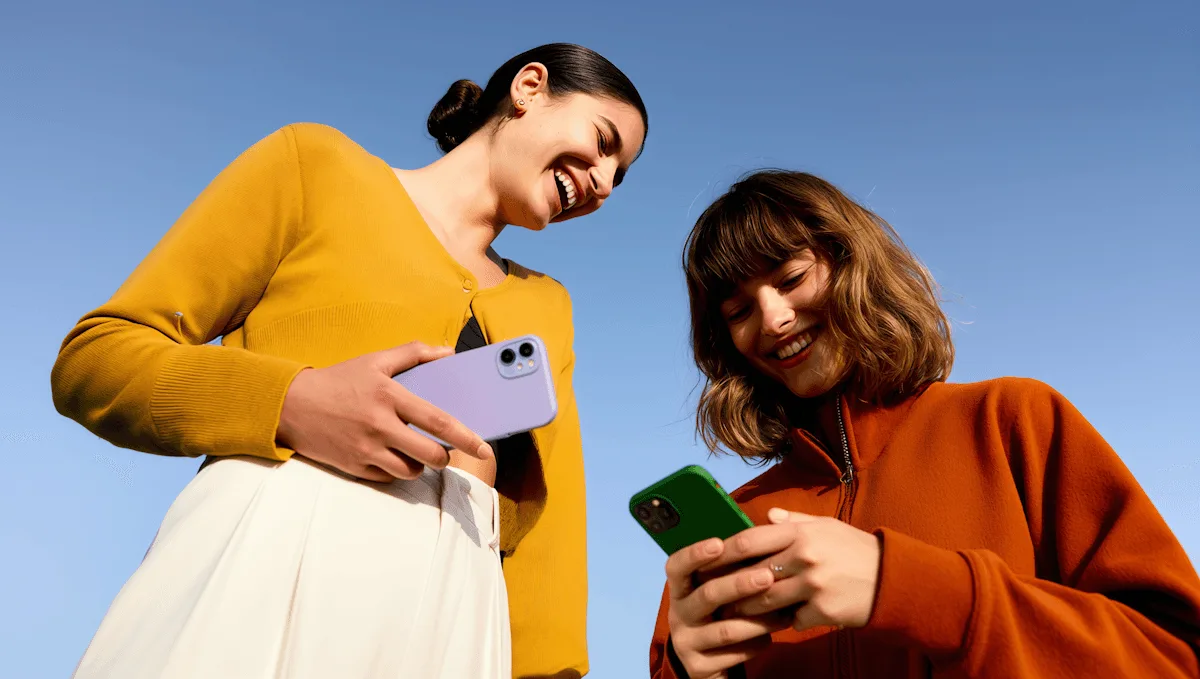Dos mujeres sonríen con sus teléfonos en la mano contra un cielo azul
