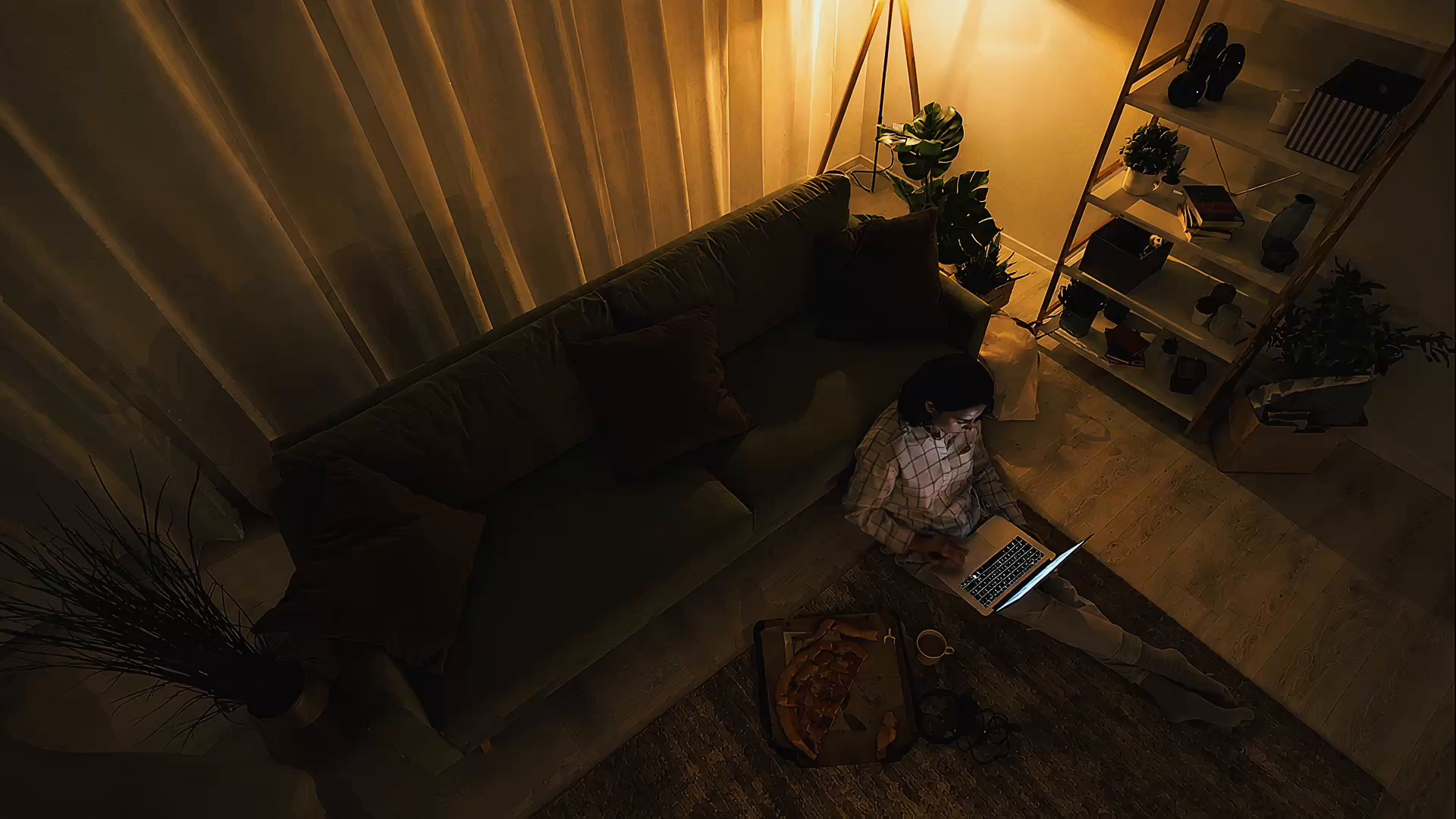 Un living a oscuras visto desde arriba, con una persona sentada en el piso contra el sillón trabajando en su notebook, una pizza a medio comer al lado y una lámpara encendida al fondo